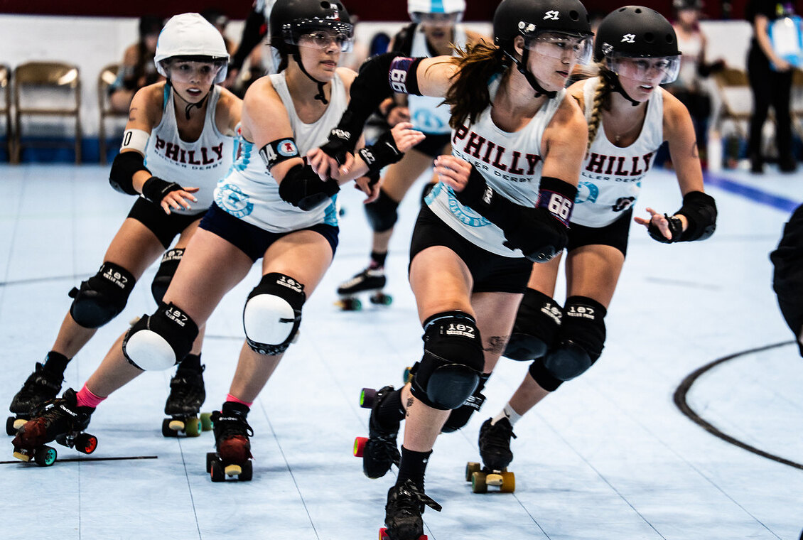 Four Philly Roller Derby skaters in helmets, mouthguards and pads, skating hard in a tight pack across a pale blue rink floor.